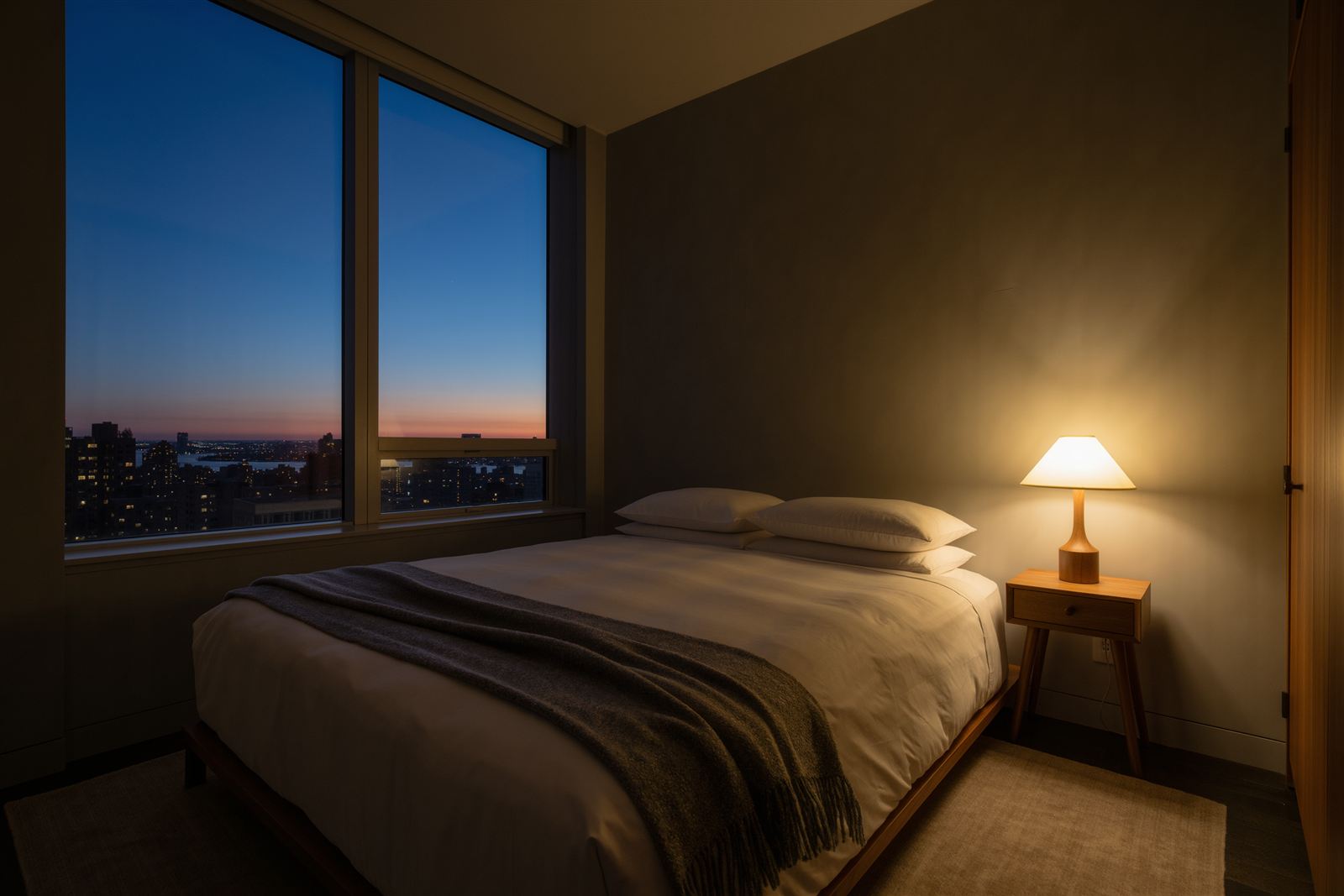 A quiet bedroom at dusk with a made bed, a lit nightstand lamp and a tall window looking out over city lights and a blue evening sky