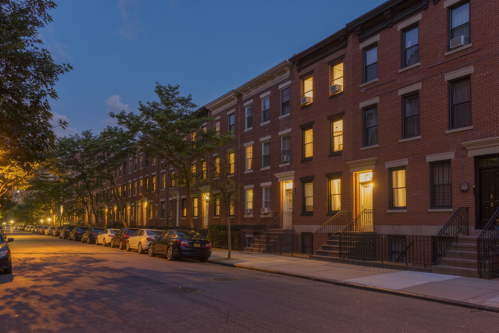 A brick low-rise Bronx street in the evening with parked cars, street trees and warmly lit windows