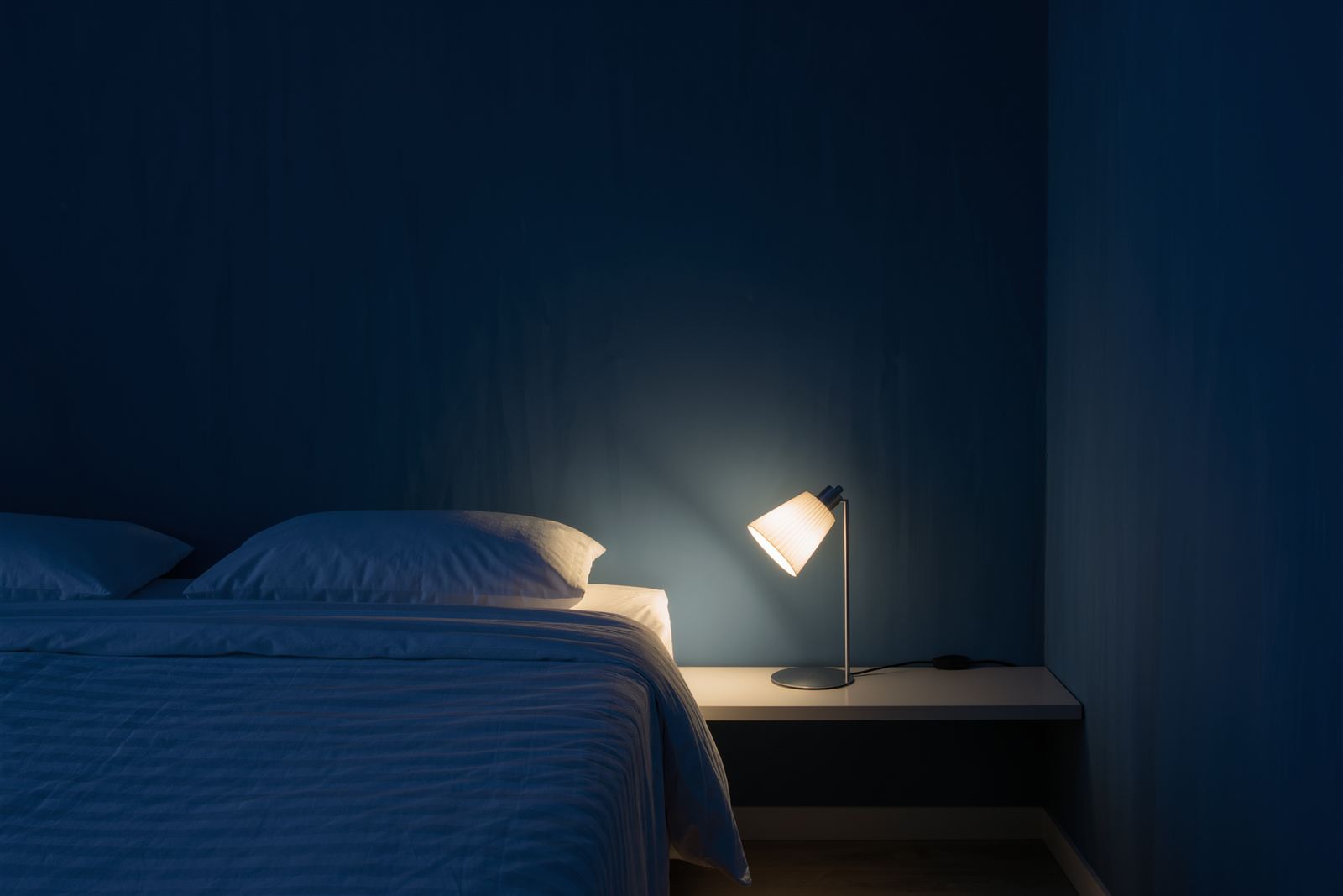 A tidy dark blue bedroom at night with a neatly made bed and a low reading lamp glowing on a narrow shelf