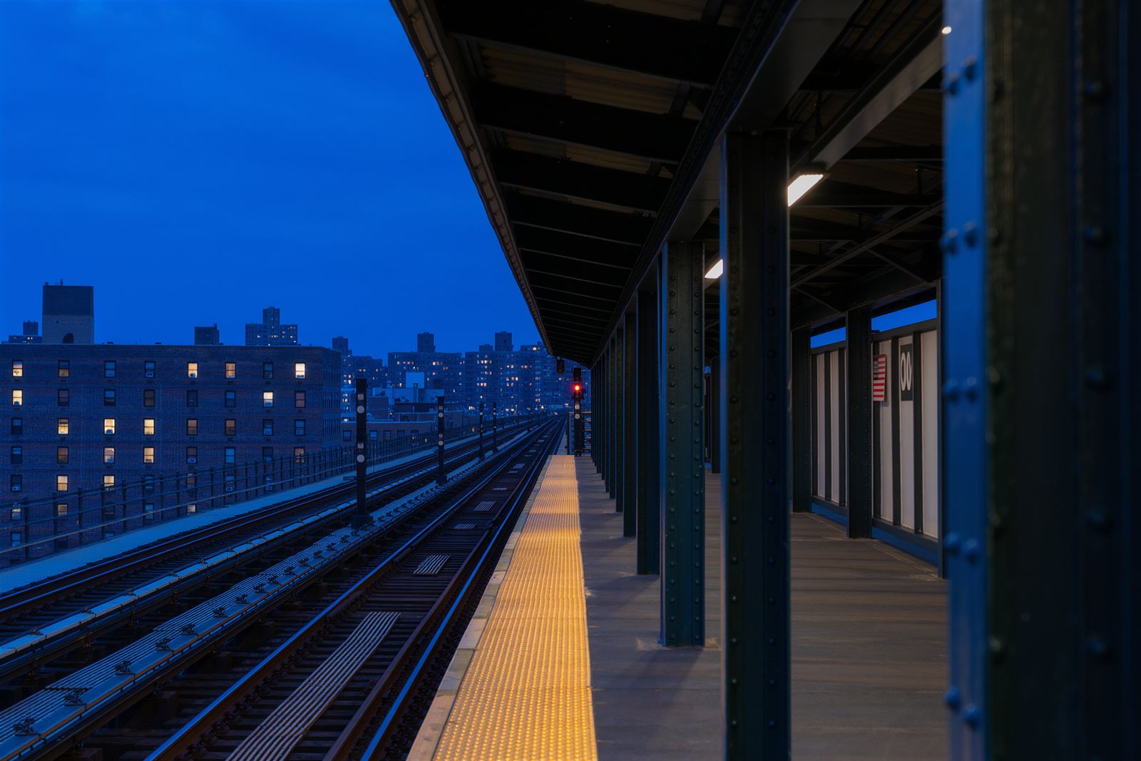 An empty elevated train platform in Queens at blue hour with steel columns, platform lights and low brick buildings beyond