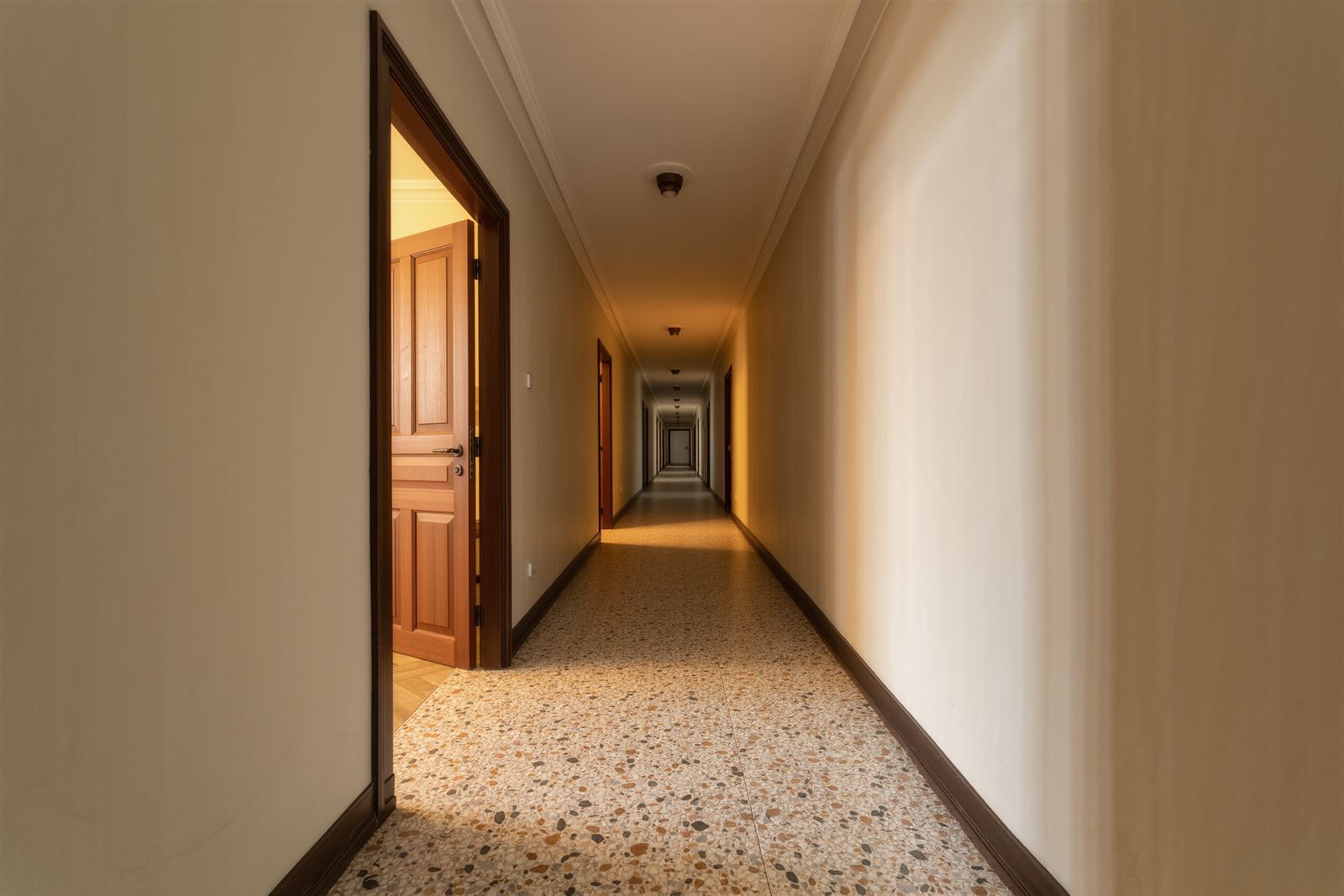 A pre-war apartment building hallway with a terrazzo floor and cream walls and one wooden door standing ajar with warm light