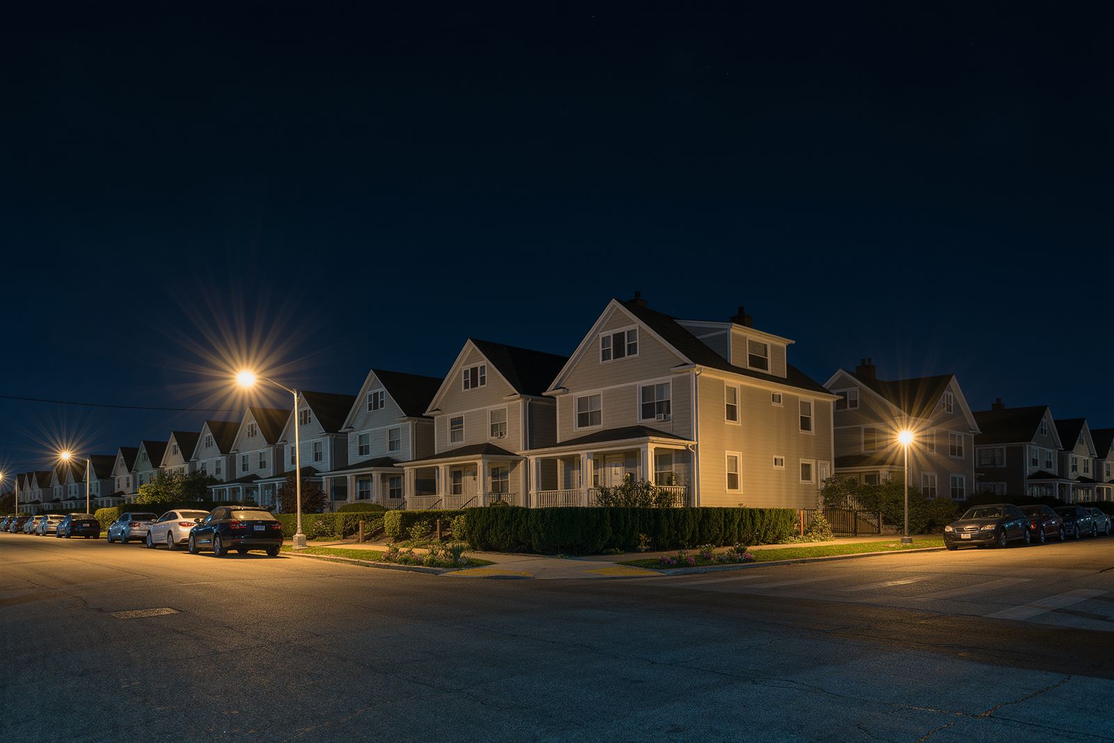 A quiet residential Bronx street at night with detached two-story houses, glowing street lamps and parked cars