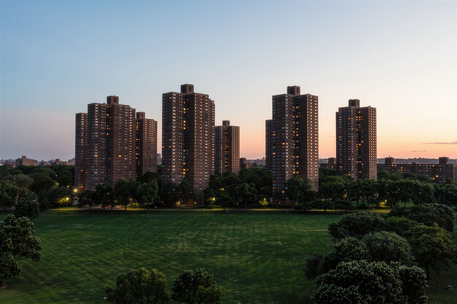 Tall brick residential towers with lit windows rising behind green lawns and trees at dusk