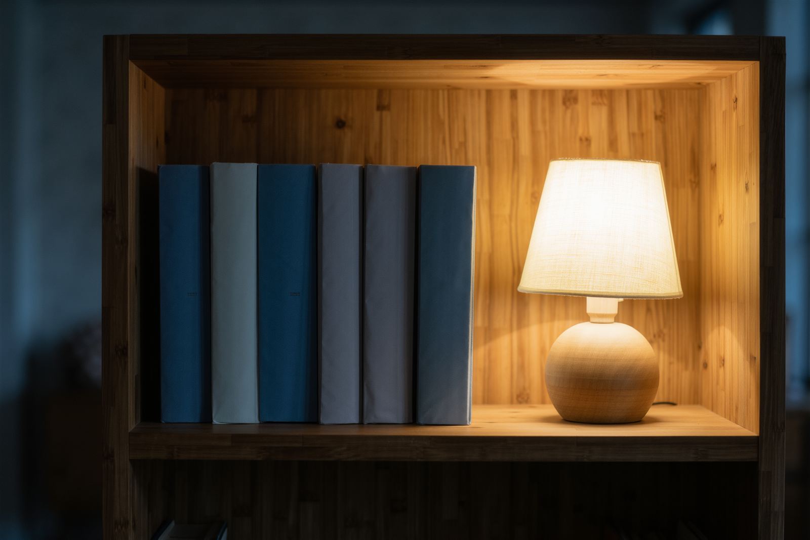 A wooden shelf with six plain blue and cream books beside a small glowing lamp in a dim room