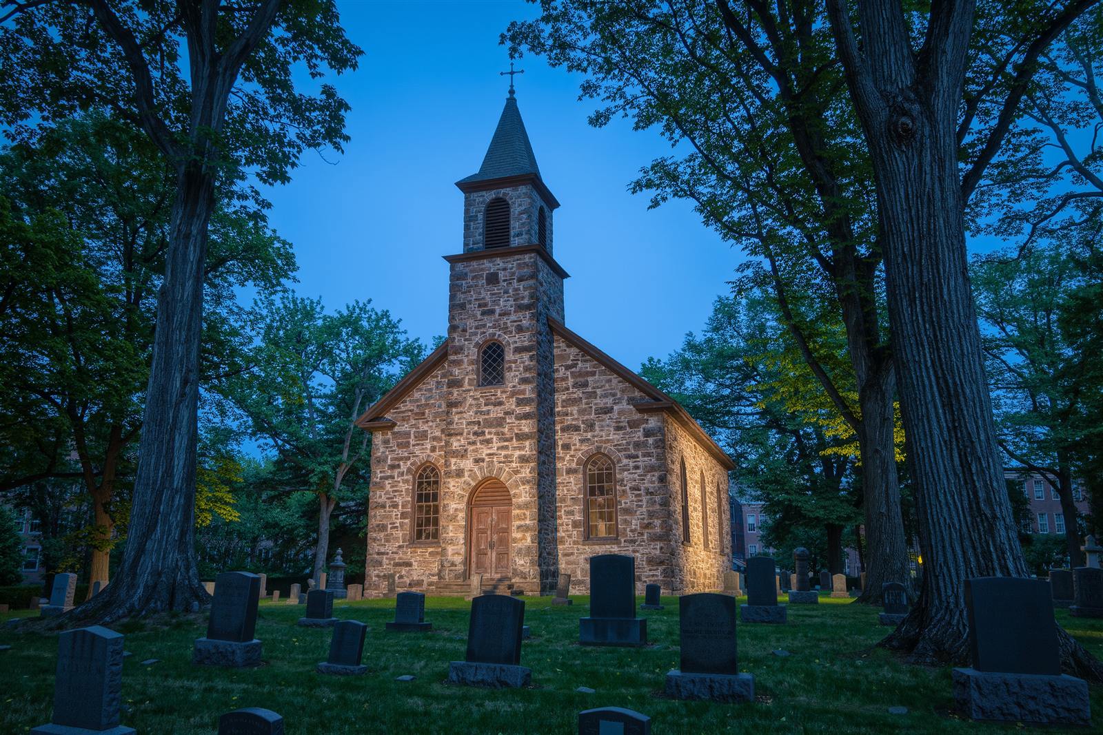 A small fieldstone church with a slate steeple among old trees and gravestones at blue hour