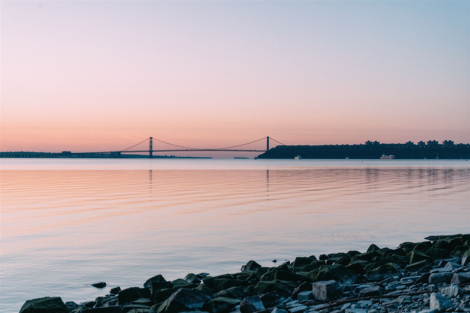 A rocky Bronx shoreline at dusk with calm pink water and a suspension bridge silhouette far across the water