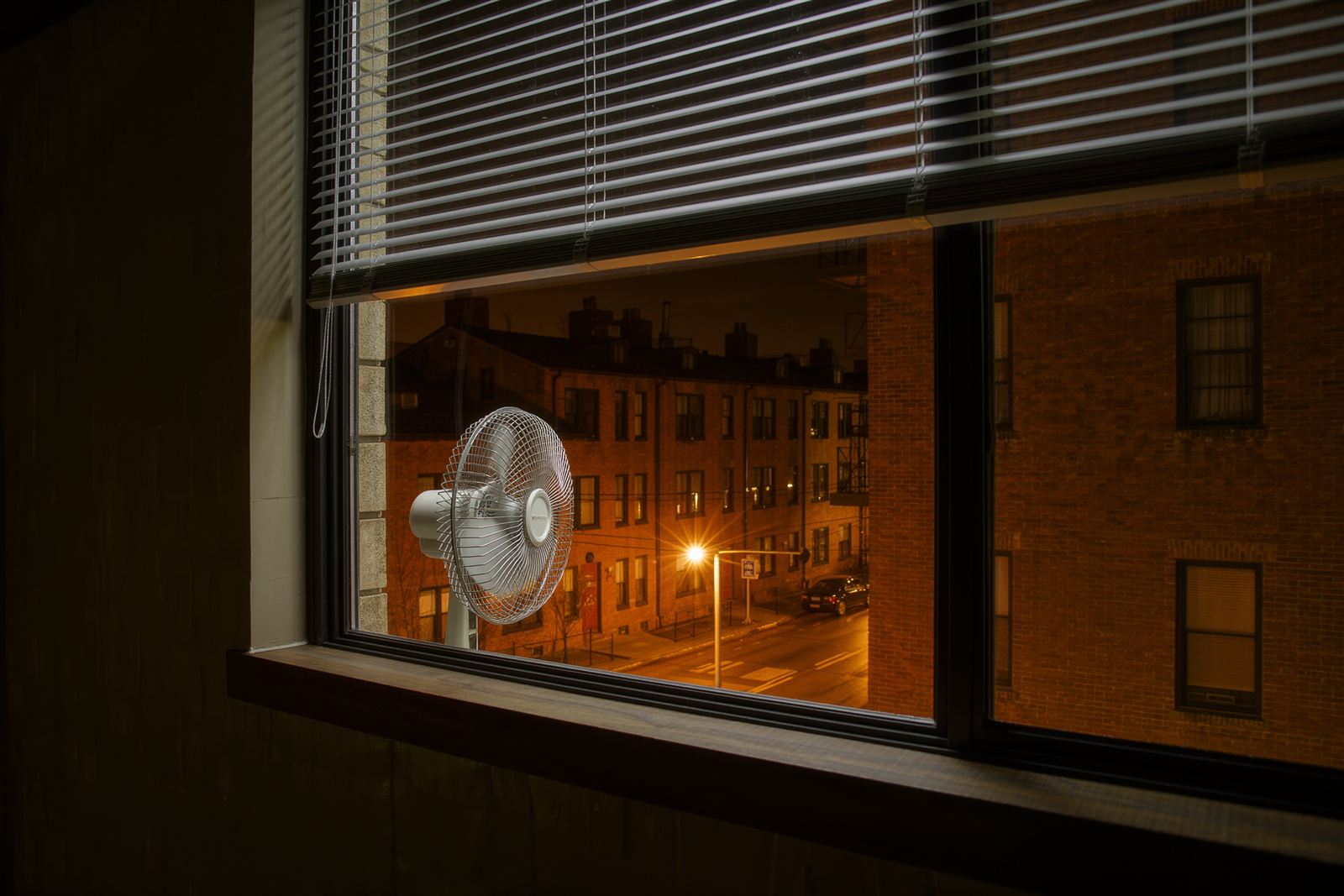 A night window with half-drawn blinds, a small fan on the sill and orange street lights over a brick street beyond