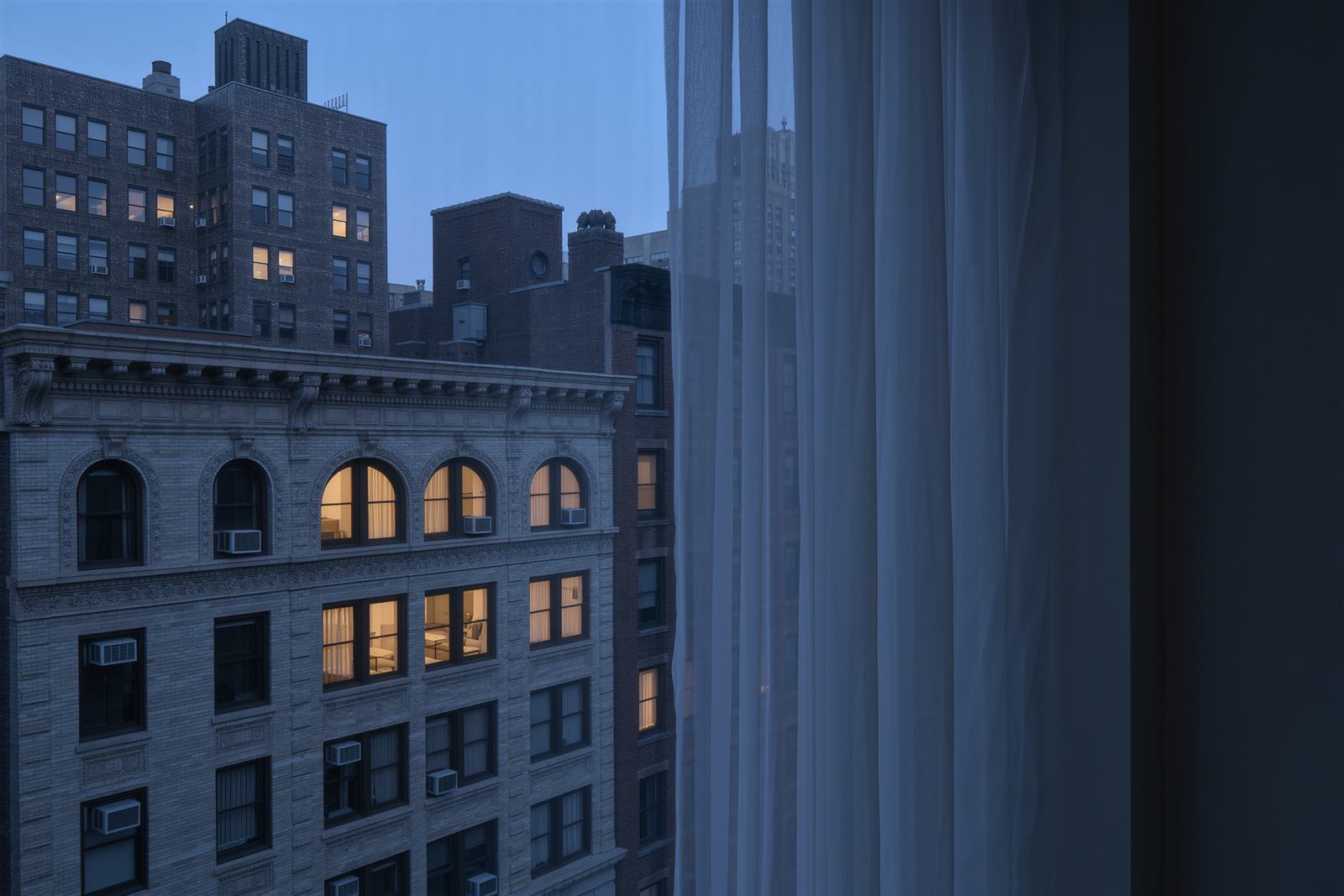A tall apartment window with a sheer curtain looking out at limestone and brick apartment buildings with lit windows at blue hour